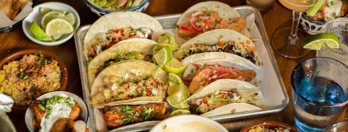 A platter of assorted tacos with lime wedges, surrounded by sides and drinks on a wooden table.