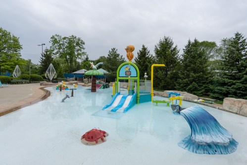 A colorful water play area with slides, water features, and playful decorations, surrounded by trees under a cloudy sky.