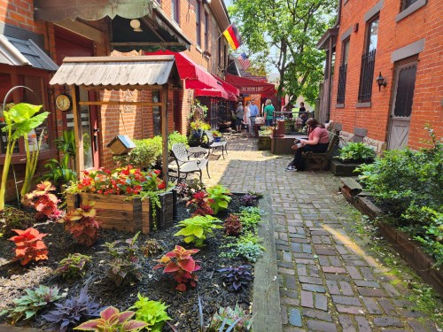 A charming alleyway lined with vibrant plants, benches, and outdoor seating at cafes, inviting visitors to relax.