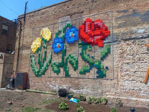 Colorful pixel art flowers on a brick wall, featuring yellow, blue, and red blooms against a textured background.