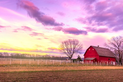 A red barn stands in a field under a colorful sunset with clouds and bare trees in the background.