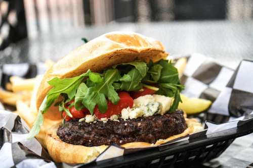 A close-up of a gourmet burger with lettuce, tomato, and blue cheese, served with fries on a checkered paper.
