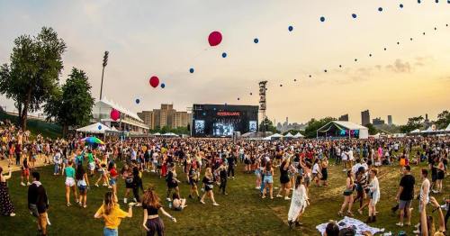 A lively outdoor music festival with a large crowd, colorful balloons, and a stage in the background during sunset.