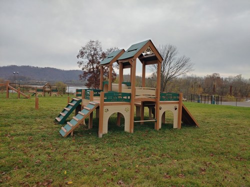 A wooden playground structure with a slide and climbing features, set in a grassy area with trees in the background.