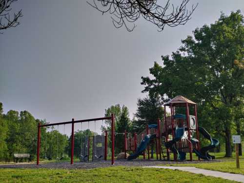 A playground with slides and swings, surrounded by trees and green grass under a cloudy sky.