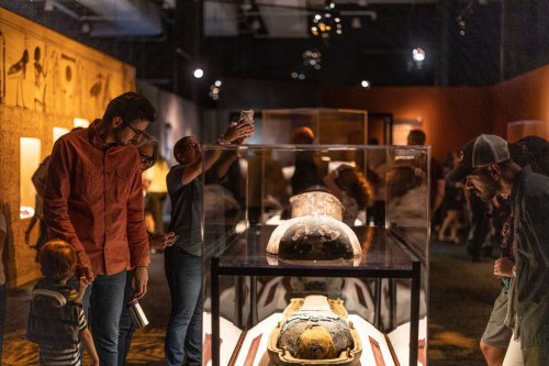 Visitors explore an exhibit featuring ancient artifacts and a displayed mummy in a dimly lit museum.