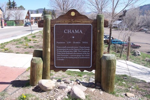 Sign for Chama, Colorado, detailing its history and population, with a scenic backdrop of trees and mountains.