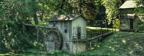 A rustic watermill with a large wooden wheel beside a serene pond, surrounded by lush greenery and trees.
