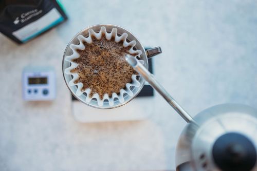 A top-down view of a coffee filter filled with grounds, with a kettle pouring water and a scale below.
