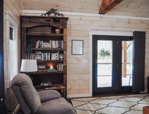 Cozy interior with a wooden bookshelf, a recliner, and double doors leading to a deck, featuring wooden walls and a rug.