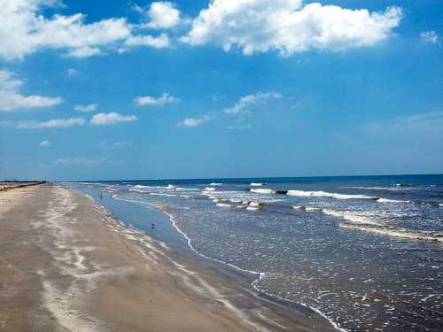 A serene beach scene with gentle waves, soft sand, and a clear blue sky dotted with clouds.