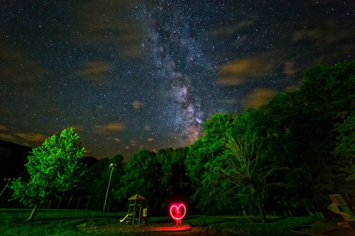 A glowing heart shape in a park under a starry sky with the Milky Way and trees in the background.