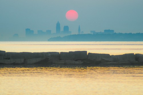 A hazy sunset over a city skyline, with a pink sun setting behind silhouettes of buildings and calm water in the foreground.