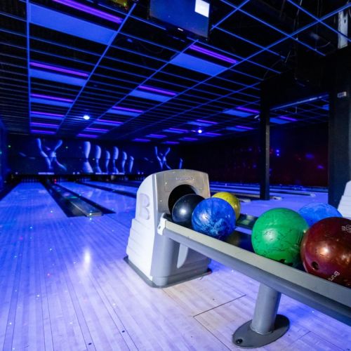 A bowling alley with colorful bowling balls on a rack, illuminated by blue and purple lights.
