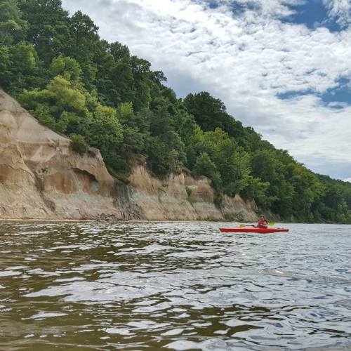 A person kayaking on calm water near a rocky, tree-lined shoreline under a partly cloudy sky.