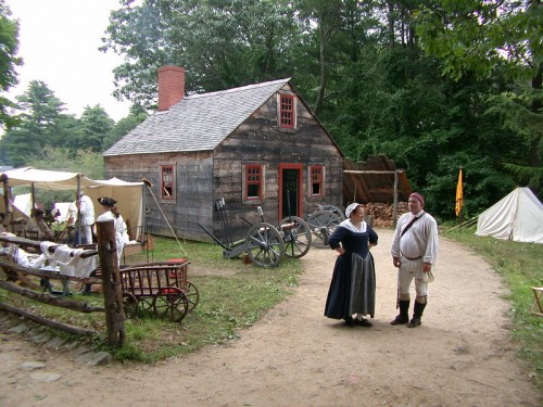 A historic scene featuring a wooden house, tents, and two people in period clothing standing on a dirt path.