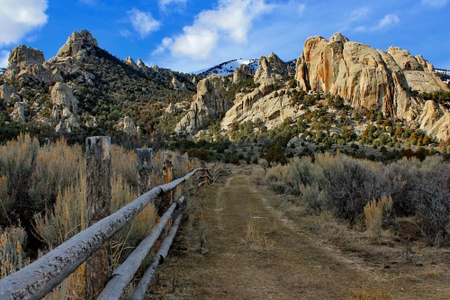 A dirt path lined with a wooden fence, leading through rocky terrain and sagebrush under a blue sky with clouds.