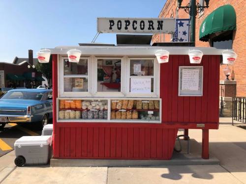 A red popcorn stand with a sign, displaying various popcorn flavors and a cooler nearby, set in a brick street.