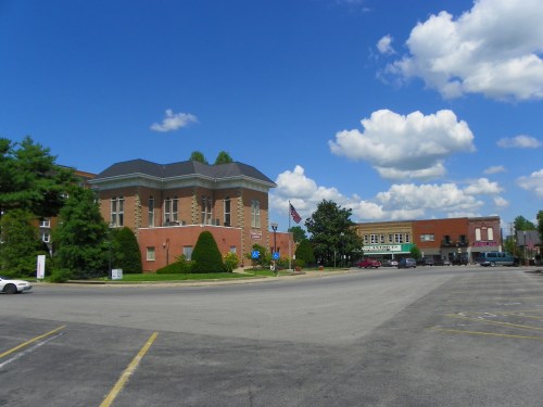 A sunny day with fluffy clouds over a small town square featuring brick buildings and well-maintained landscaping.