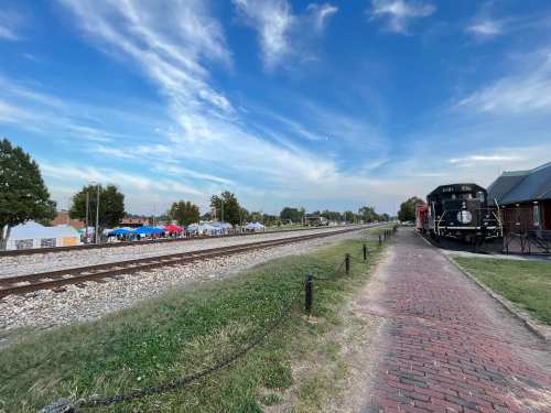 A scenic view of a railway track beside a brick path, with a vintage train and tents in the background under a blue sky.