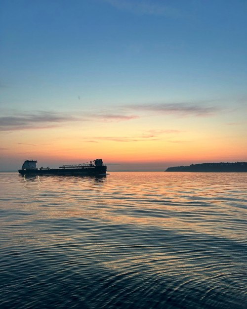 A tranquil seascape at sunset, featuring a ship on calm waters with a colorful sky and distant shoreline.