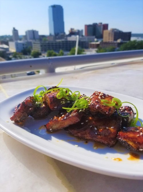 A plate of glazed ribs garnished with green onions, with a city skyline in the background under a clear blue sky.