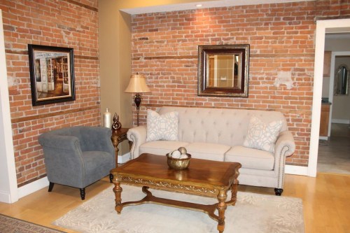 Cozy living room with exposed brick walls, a beige sofa, gray armchair, wooden coffee table, and decorative accents.