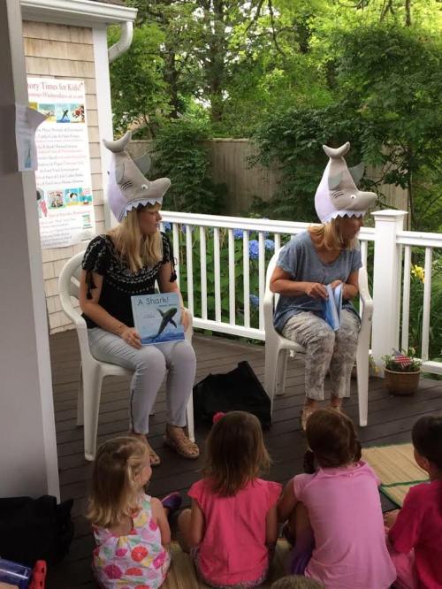 Two women wearing shark hats sit on chairs, reading to a group of children on a porch surrounded by greenery.