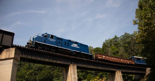 A blue locomotive crosses a bridge, pulling a train with "POTOMAC EAGLE" written on the side against a clear sky.