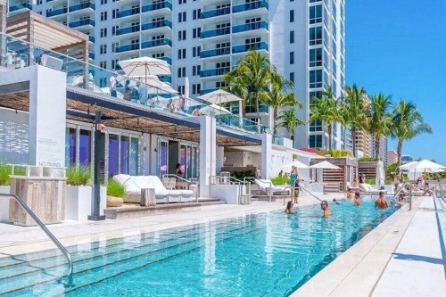 A sunny pool area with lounge chairs, umbrellas, and people enjoying the water, surrounded by modern buildings and palm trees.