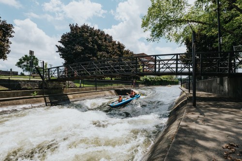 A kayak navigates a river with rapids under a bridge, surrounded by trees and a sunny sky.
