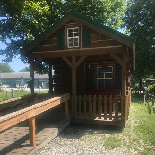 A small wooden cabin with a porch, surrounded by trees and a grassy area, near a pool in the background.
