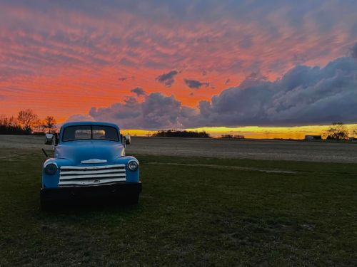 A vintage blue truck parked on grass, with a vibrant sunset and colorful clouds in the background.