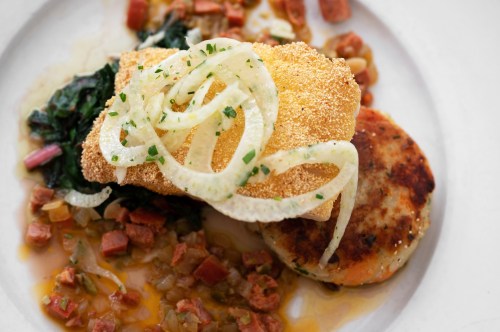 A plate of golden-fried fish topped with onions, served with sautéed greens and a savory potato cake.