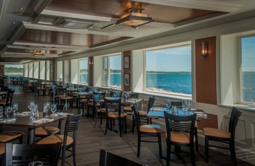 Bright restaurant interior with wooden tables and chairs, large windows overlooking a scenic waterfront view.
