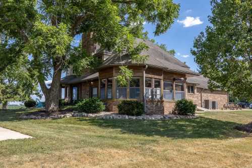 A brick house with a large porch, surrounded by trees and a grassy yard under a blue sky with fluffy clouds.