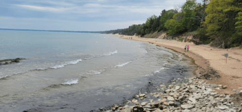 A sandy beach with gentle waves, surrounded by trees and a rocky shoreline, under a cloudy sky. People walk in the distance.