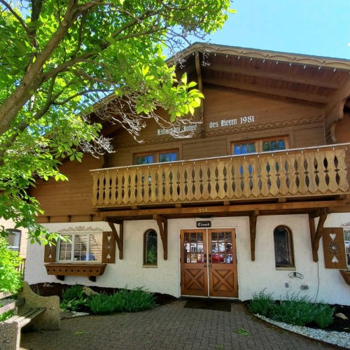 A charming wooden chalet with a balcony, surrounded by greenery and featuring decorative elements on the facade.
