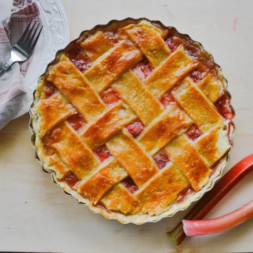 A freshly baked lattice-top pie filled with red fruit, placed on a light wooden surface with a fork and napkin nearby.