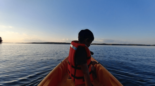 A child in a life jacket sits in a kayak on calm water, with a clear blue sky and distant trees in the background.