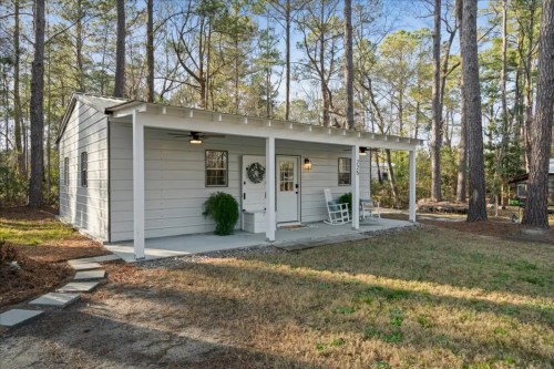 A small, charming white cottage with a porch, surrounded by trees and a grassy area.