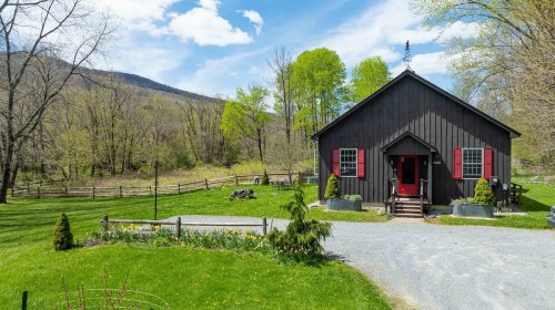 A charming black house with red accents surrounded by greenery and mountains under a blue sky with fluffy clouds.