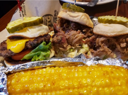Two mini sandwiches with pickles, lettuce, tomato, and cheese, alongside a grilled corn on the cob.