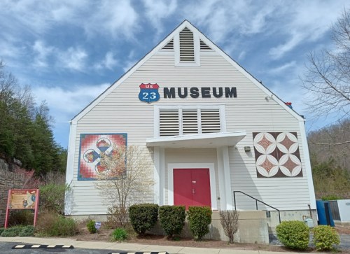 Exterior of a museum building with a triangular roof, featuring colorful murals and a red door. Clear blue sky above.