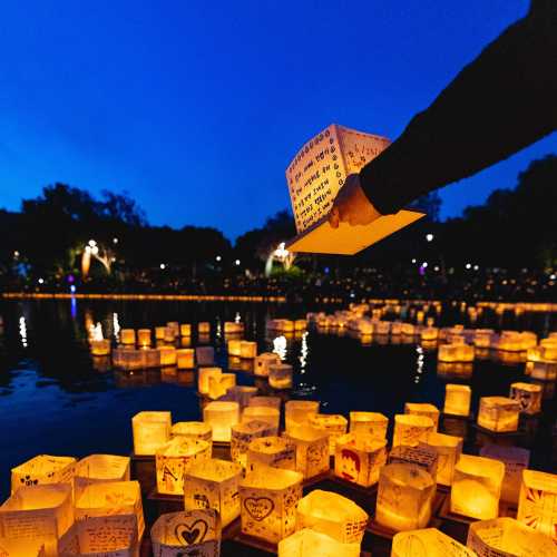 A hand holds a lit lantern above a lake filled with glowing paper lanterns at dusk.