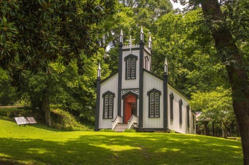 A quaint, white church with black trim and spires, surrounded by lush green trees and grass.