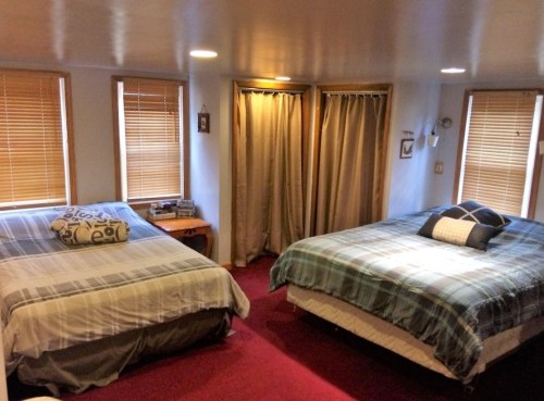 Cozy bedroom with two beds, wooden blinds, and warm lighting, featuring a red carpet and decorative pillows.