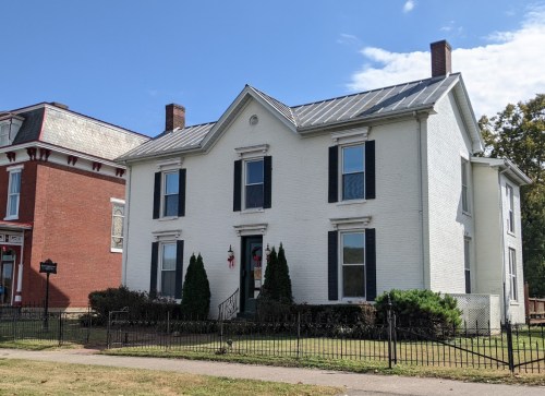 A white two-story house with black shutters, a metal roof, and a small front yard, surrounded by a black fence.