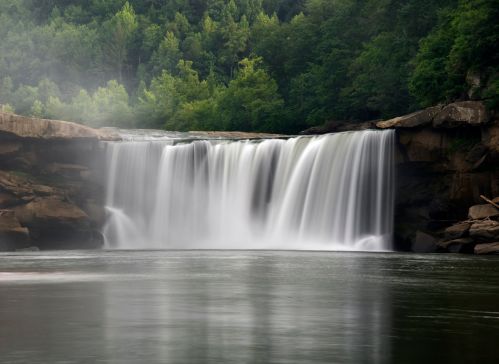 A serene waterfall cascades over rocks into a calm river, surrounded by lush green trees and misty air.