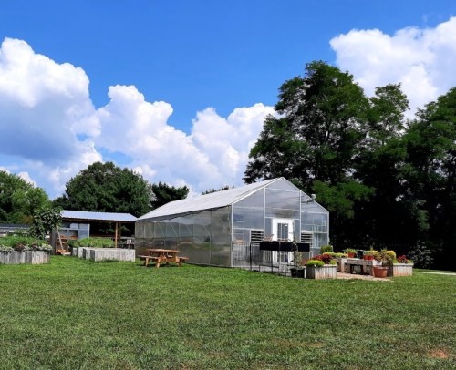 A large greenhouse surrounded by garden beds and picnic tables under a blue sky with fluffy clouds.